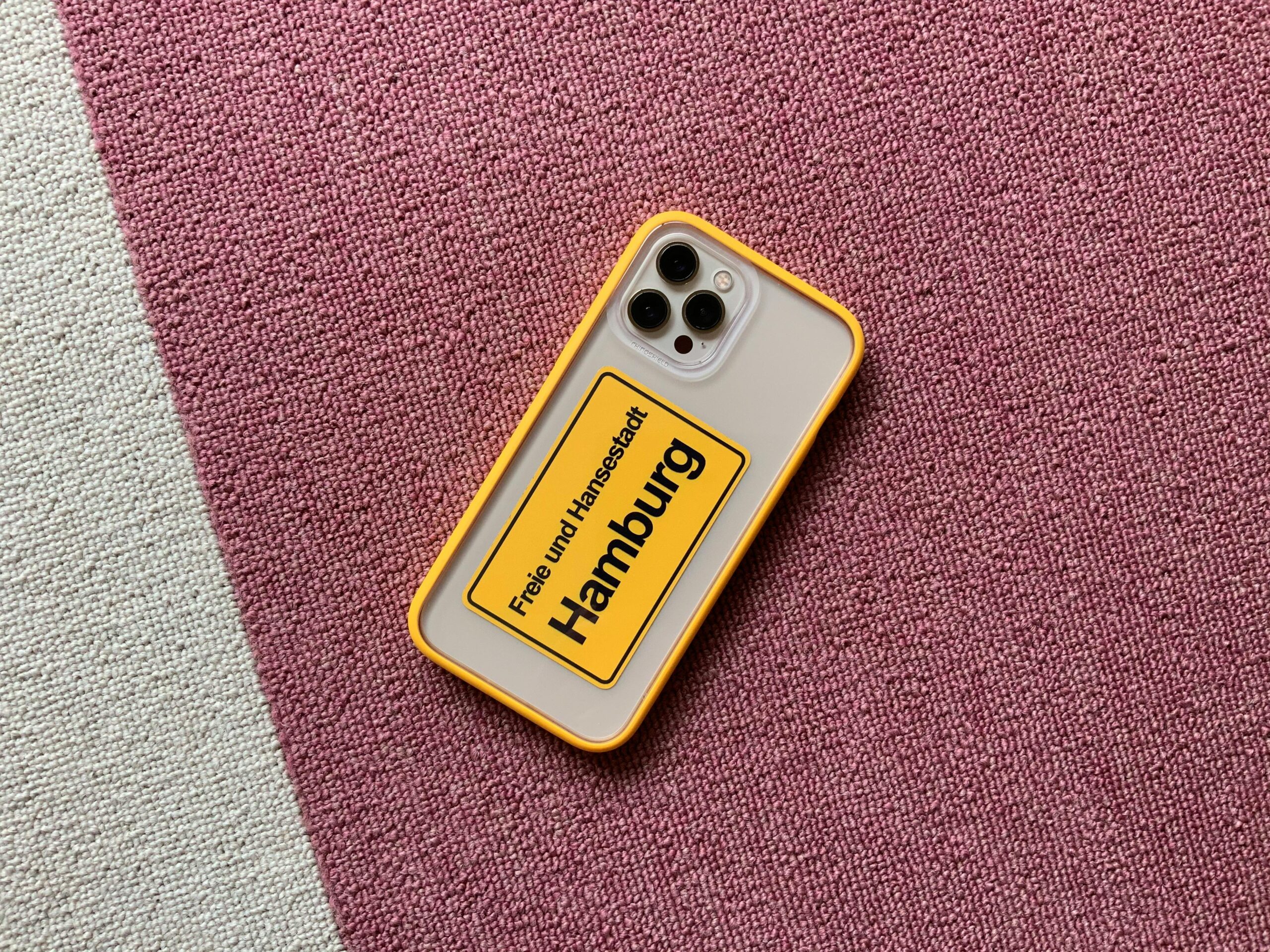Smartphone with vibrant yellow case displaying 'Hamburg' on a textured pink carpet.